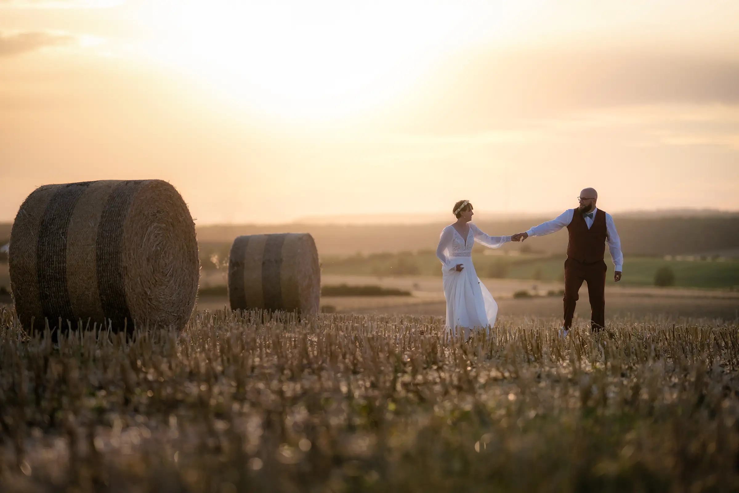 Les mariés marchent main dans la main entre des bottes de paille au coucher du soleil lors d'une séance Day After à Nancy